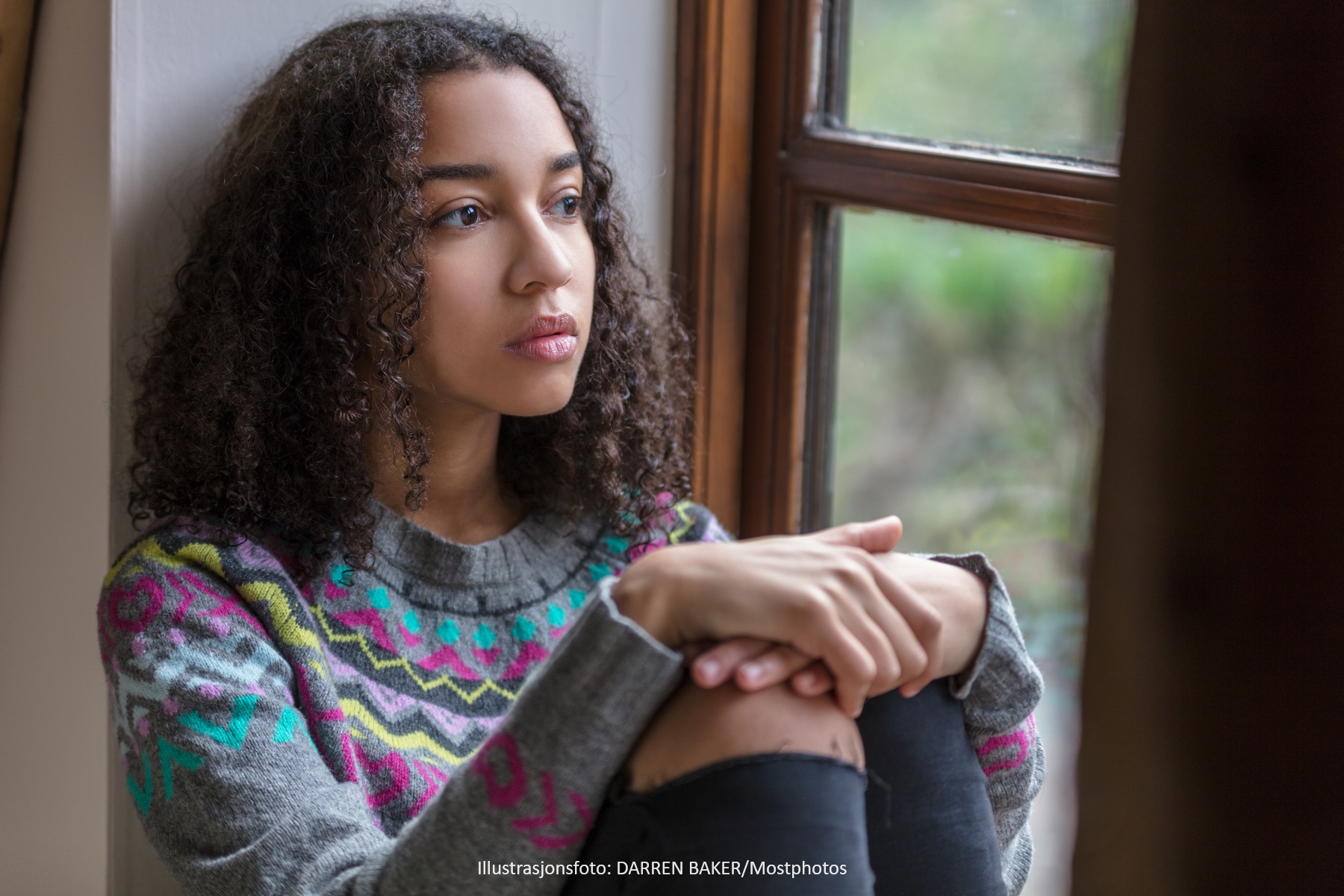 Illustration photo. A teenage girl sits alone by a window, looking out.